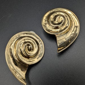 Peut inclure: Une paire de boucles d'oreilles dorées en forme de coquilles de nautile stylisées. Chaque boucle d'oreille présente une spirale centrale proéminente avec une finition métallique lisse et réfléchissante et une extrémité pointue effilée.
