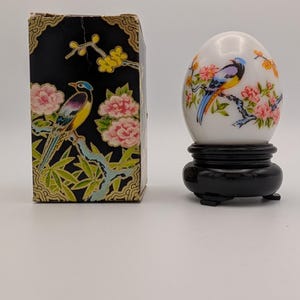 Op de afbeelding: Een wit eivormig ornament met een kleurrijk vogel- en bloemmotief, rustend op een zwarte ronde standaard. Daarnaast staat een rechthoekige zwarte doos versierd met een bijpassende illustratie van een vogel op een bloeiende tak.