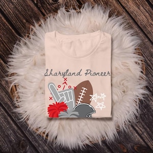 May include: A beige t-shirt with the text "Maryland Pioneer" printed on the front. The shirt features a graphic design of a hand making a number one gesture, a football, a pom-pom, and a star.