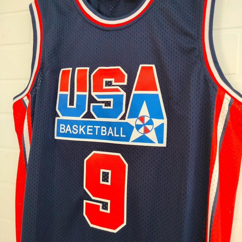 May include: A blue basketball jersey with red and white trim. The jersey has the word "USA" in large red, white, and blue letters with the word "BASKETBALL" underneath. The number "9" is on the front of the jersey in red with a white outline.