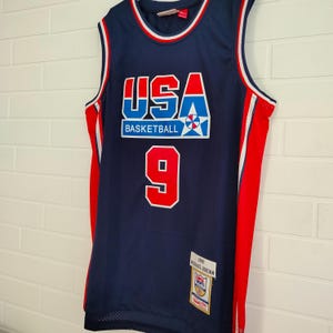 May include: A blue basketball jersey with red and white trim. The jersey has the word "USA" in large letters with a star and the words "Basketball" underneath. The number "9" is on the front of the jersey. The jersey has a tag that says "1992 Michael Jordan".