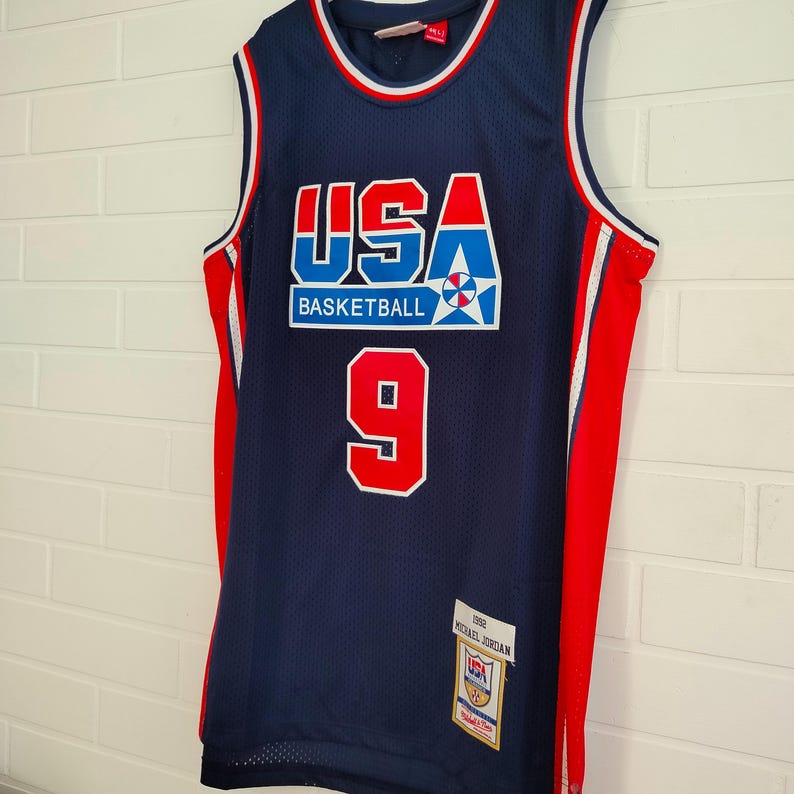 May include: A blue basketball jersey with red and white trim. The jersey has the word "USA" in large letters with a star and the words "Basketball" underneath. The number "9" is on the front of the jersey. The jersey has a tag that says "1992 Michael Jordan".