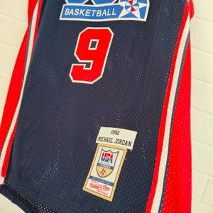 May include: A blue basketball jersey with red and white stripes. The jersey has the number "9" on the front and the text "1992 MICHAEL JORDAN" on the bottom. The jersey also has a patch with the text "USA BASKETBALL CLASSICS" and "AUTHENTIC Mitchell & Ness PHILADELPHIA, PA."