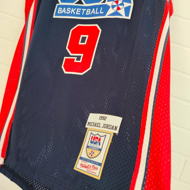 May include: A blue basketball jersey with red and white stripes. The jersey has the number "9" on the front and the text "1992 MICHAEL JORDAN" on the bottom. The jersey also has a patch with the text "USA BASKETBALL CLASSICS" and "AUTHENTIC Mitchell & Ness PHILADELPHIA, PA."
