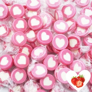 Puede incluir: Caramelos de corazón rosa y blanco envueltos en plástico transparente. Los caramelos están dispuestos en una pila, con los corazones hacia arriba.