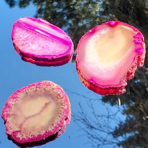 May include: Three polished agate slices with concentric bands of pink and white, displayed on a reflective surface. The natural stone pieces have a vibrant color palette and unique patterns, showcasing their geological beauty.
