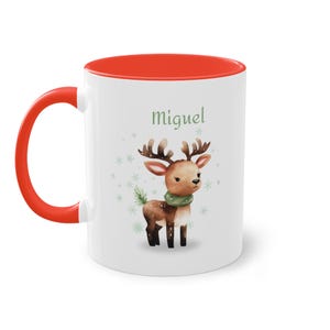 Op de afbeelding: Witte keramische mok met een rode binnenkant en handvat. De mok heeft een aquarel illustratie van een rendier met een groene sjaal, met de naam "Miguel" in groene letters. Sneeuwvlok accenten maken het ontwerp af.