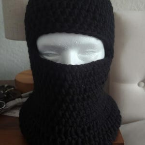 May include: A black, crocheted balaclava, displayed on a white mannequin head. The head covering features a knitted design with an opening for the eyes and mouth. The balaclava is made of a thick, dark yarn.