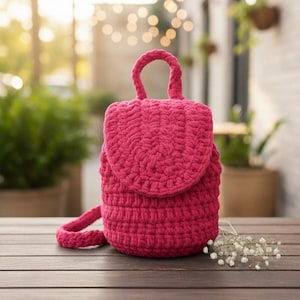 Puede incluir: Mochila de ganchillo rosa intenso hecha a mano, con cierre de solapa y asa superior. La bolsa está hecha de hilo grueso, y la textura es visible. La mochila está sobre una mesa de madera, con un fondo borroso de vegetación.