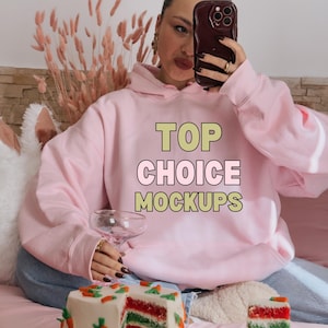 Op de afbeelding: Een roze hoodie met de tekst "TOP CHOICE MOCKUPS" op de voorkant. De hoodie wordt gedragen door een persoon die op een bank zit met een stuk gelaagde cake op een bord voor haar.