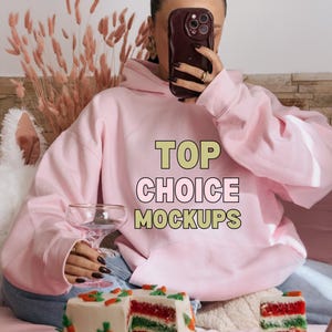 Puede incluir: Una sudadera rosa con el texto "TOP CHOICE MOCKUPS" en la parte delantera. La sudadera la lleva puesta una persona que sostiene un teléfono y está sentada en un sofá con un pastel y una copa de champán.