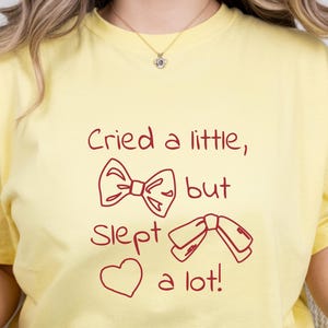 May include: A light yellow t-shirt with the text "Cried a little, but slept a lot!" in red. The design includes a bow, a heart, and a bow tie. The shirt is a casual, comfortable style.