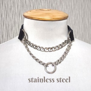 Op de afbeelding: Een zwarte choker-halsketting met twee zilverkleurige roestvrijstalen kettingen die aan beide zijden aan ronde ringen zijn bevestigd. De onderste ketting hangt in een gedrapeerde stijl en is verbonden met een centrale ring. De tekst stainless steel is zichtbaar.