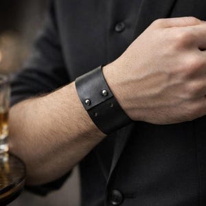 May include: A black leather cuff bracelet with silver rivets. The wide band wraps around the wrist and fastens with two rivets. The bracelet is worn on a person's arm, with a dark background.