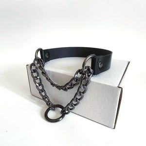 Peut inclure: Collier ras du cou en cuir noir avec chaînes en métal canon de fusil et un grand anneau. Le choker a un design simple avec une fermeture à boucle. Les chaînes pendent en couches, ajoutant un look audacieux et audacieux. Le collier est présenté sur une surface blanche.