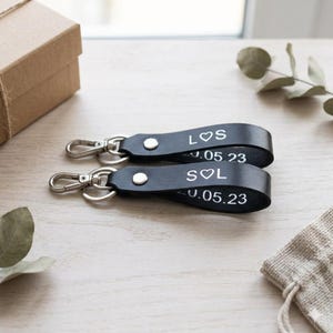 May include: Two black leather keychains with silver-tone hardware. Each keychain has white text that reads "LOS ♡ S" and "S ♡ L" with the date "0.05.23". The keychains are on a light wood surface.
