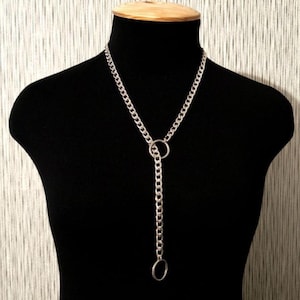 May include: Silver-toned chain necklace with a pendant. The necklace features a thick chain that forms a loop around the neck, with a longer chain and a ring pendant hanging down the front. The jewelry is displayed on a black mannequin.