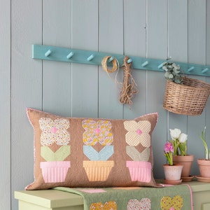 May include: A rectangular brown pillow with a floral design. The pillow features three appliqued flower pot designs with colorful flowers. The background is a light brown color. The pillow is resting on a green table runner with a matching floral pattern.