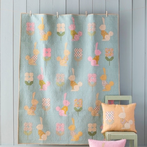 May include: A light blue quilt with a repeating pattern of bunnies and flowers in pastel colors. Two matching throw pillows are on a small green chair. The quilt is hanging on a clothesline.