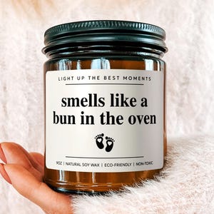 May include: A candle in a glass jar with a black lid. The label reads "smells like a bun in the oven" with baby footprints. The candle is held in front of a soft, pink sweater.