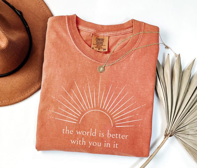 Op de afbeelding: Een koraalkleurig t-shirt met een wit zonnestraalontwerp en de tekst "the world is better with you in it." Een gouden ketting met een hanger ligt op het shirt. Een bruine hoed en gedroogde palmbladeren staan ook op de foto.