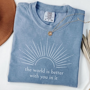 Op de afbeelding: Een blauw Comfort Colors T-shirt met een wit zonnestraalontwerp en de tekst "the world is better with you in it." Een gouden ketting met een hanger ligt op het shirt. Het shirt is maat small.