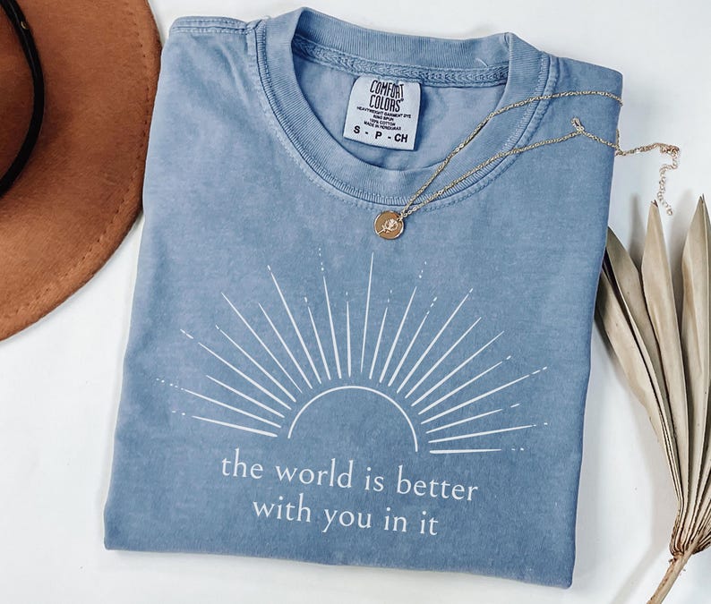 Op de afbeelding: Een blauw Comfort Colors T-shirt met een wit zonnestraalontwerp en de tekst "the world is better with you in it." Een gouden ketting met een hanger ligt op het shirt. Het shirt is maat small.