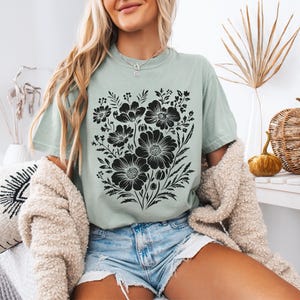 May include: A light green t-shirt featuring a large black floral design. The graphic includes various flower shapes and foliage. The shirt is paired with denim shorts and a cream-colored cardigan. The overall style is casual and relaxed.