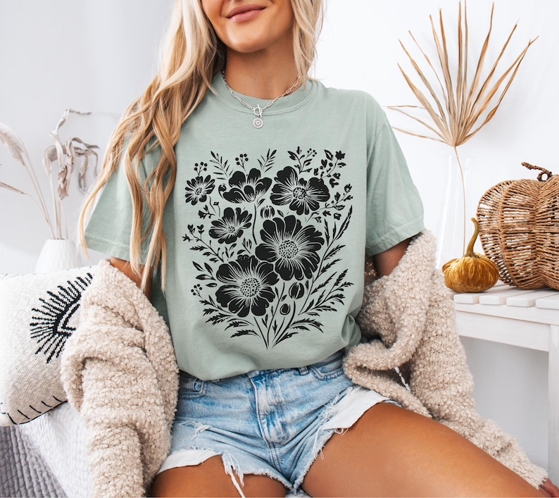 May include: A light green t-shirt featuring a large black floral design. The graphic includes various flower shapes and foliage. The shirt is paired with denim shorts and a cream-colored cardigan. The overall style is casual and relaxed.