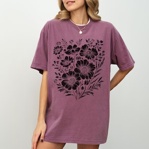 May include: A mauve-colored t-shirt featuring a large black floral design. The graphic includes various flower types and foliage, creating a detailed botanical illustration. The shirt has short sleeves and a relaxed fit, suitable for casual wear.