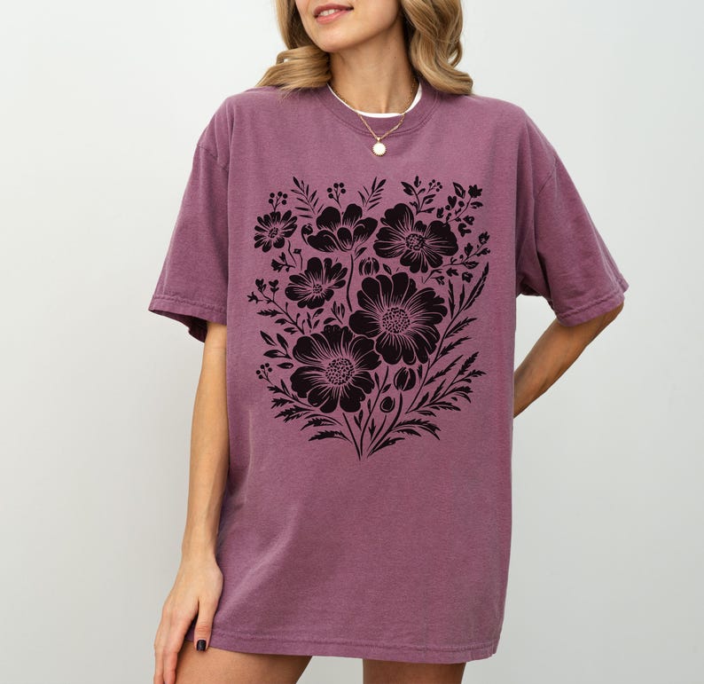 May include: A mauve-colored t-shirt featuring a large black floral design. The graphic includes various flower types and foliage, creating a detailed botanical illustration. The shirt has short sleeves and a relaxed fit, suitable for casual wear.