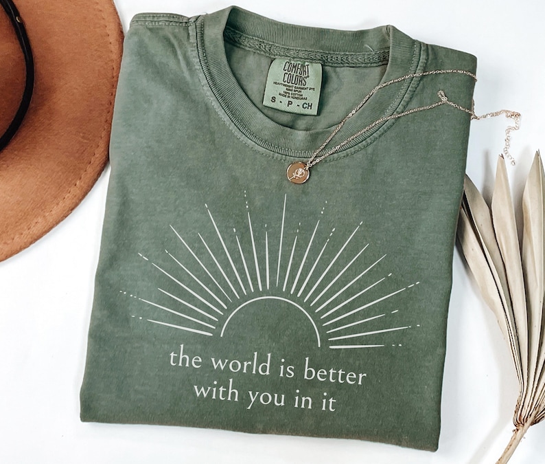 Op de afbeelding: Olijfgroen t-shirt met een witte zonnestraal en de tekst "the world is better with you in it." Een gouden ketting met een kleine hanger ligt op het shirt. Een bruine hoed en een gedroogde plant staan op de achtergrond.