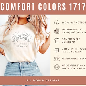 Op de afbeelding: Cr&egrave;mekleurig unisex T-shirt met de tekst "the world is better with you in it" en een zonnestraalontwerp. Het shirt is gemaakt van 100% USA katoen, is van gemiddeld gewicht en heeft een verweerde vintage look. De print is direct en zal niet afbladderen.