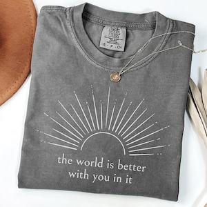 Op de afbeelding: Grijs Comfort Colors T-shirt met een wit zonnestraalontwerp en de tekst "the world is better with you in it." Een gouden ketting en een bruine hoed staan ook op de foto.