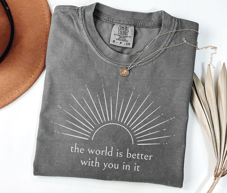 Op de afbeelding: Grijs Comfort Colors T-shirt met een wit zonnestraalontwerp en de tekst "the world is better with you in it." Een gouden ketting en een bruine hoed staan ook op de foto.