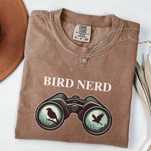 May include: A brown Comfort Colors t-shirt with the words "BIRD NERD" in white. Below the text is a graphic of binoculars with birds inside the lenses. A silver necklace with a bird pendant and a brown hat are also in the image.