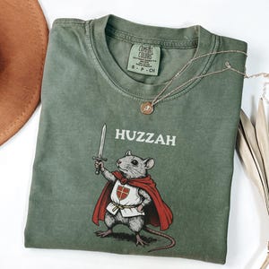 May include: Sage green t-shirt with the word "Huzzah" above a cartoon mouse holding a sword and wearing a red cape. A gold necklace with a pendant rests on the shirt. The shirt has a Comfort Colors tag.