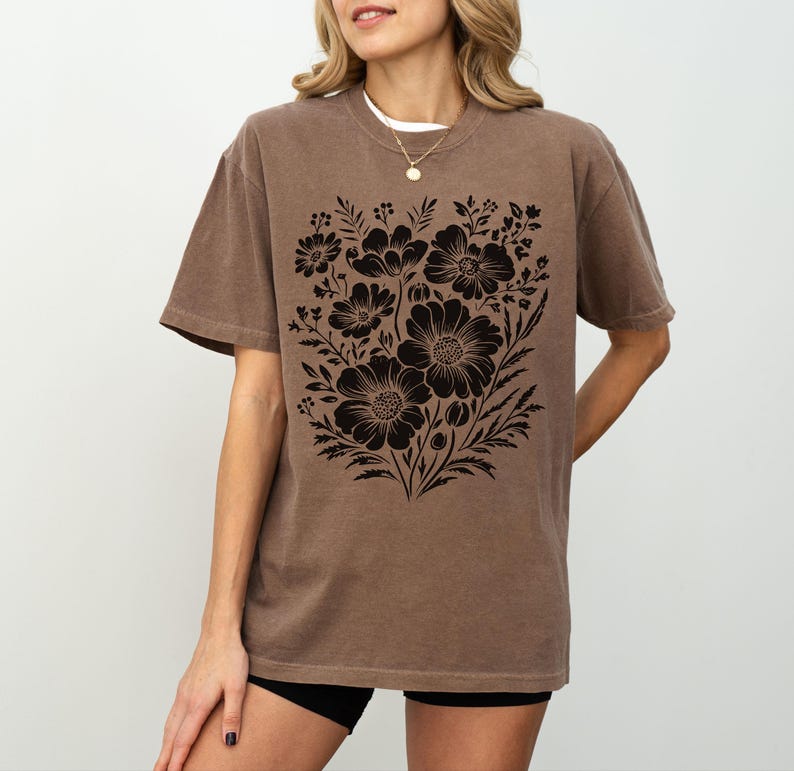 May include: Brown t-shirt featuring a black floral design. The graphic showcases a detailed arrangement of flowers and leaves. The shirt has short sleeves and a relaxed fit, suitable for casual wear.