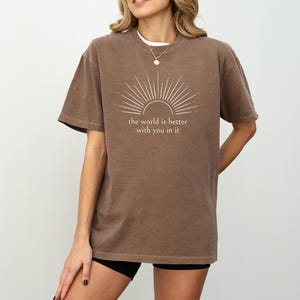 Op de afbeelding: Bruin t-shirt met de witte tekst "the world is better with you in it". De afbeelding boven de tekst is een sunburst-ontwerp. Het shirt heeft korte mouwen en een ronde hals.