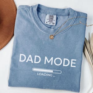 May include: A blue Comfort Colors t-shirt with the words "DAD MODE" in white block letters. Below the text is a loading bar graphic. A gold necklace and a brown hat are also in the image.