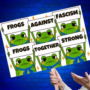 May include: A sign with cartoon frogs holding placards. The signs read "FROGS AGAINST FASCISM STRONG TOGETHER." The frogs are green with blue bow ties, set against a blue brick wall. A hand holds the sign.