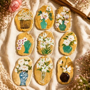 Peut inclure: Neuf plaques en argile de forme ovale avec des arrangements floraux peints à la main. Les motifs présentent des fleurs blanches dans divers vases, notamment des récipients bleu sarcelle, à motifs bleus et brun foncé, sur un fond terreux.