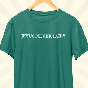 Könnte beinhalten: Ein teal-farbenes T-Shirt mit dem weißen Aufdruck "JESUS NEVER FAILS". Das Shirt hängt an einem Holzbügel vor einem cremefarbenen Hintergrund mit hellbraunen Streifen.
