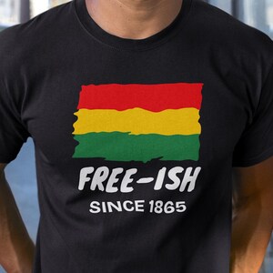 Op de afbeelding: Een zwart t-shirt met een afbeelding van een rood, geel en groen horizontaal gestreepte vlag met een rafelige rand. Onder de vlag staat de witte tekst "FREE-ISH" in een vetgedrukt lettertype, gevolgd door de witte tekst "SINCE 1865" in kleiner lettertype.