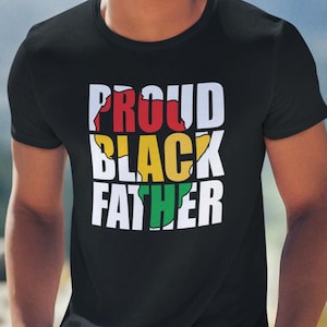 May include: Black t-shirt with the text "PROUD BLACK FATHER" in white, red, yellow, and green. The shirt is worn by a person with a mountain backdrop.