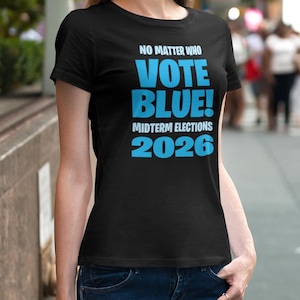Op de afbeelding: Zwart T-shirt met de tekst "NO MATTER WHO VOTE BLUE! MIDTERM ELECTIONS 2026" in helderblauw. Het T-shirt heeft een ronde hals en wordt door een persoon gedragen.
