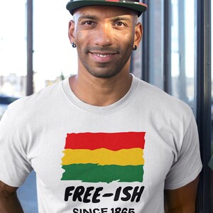 Op de afbeelding: Een wit T-shirt met korte mouwen en een afbeelding van een rood, geel en groen horizontaal gestreepte vlag met een verweerde textuur. Onder de vlag staat de tekst "FREE-ISH" in dikke zwarte letters, gevolgd door "SINCE 1865" in kleinere zwarte letters.