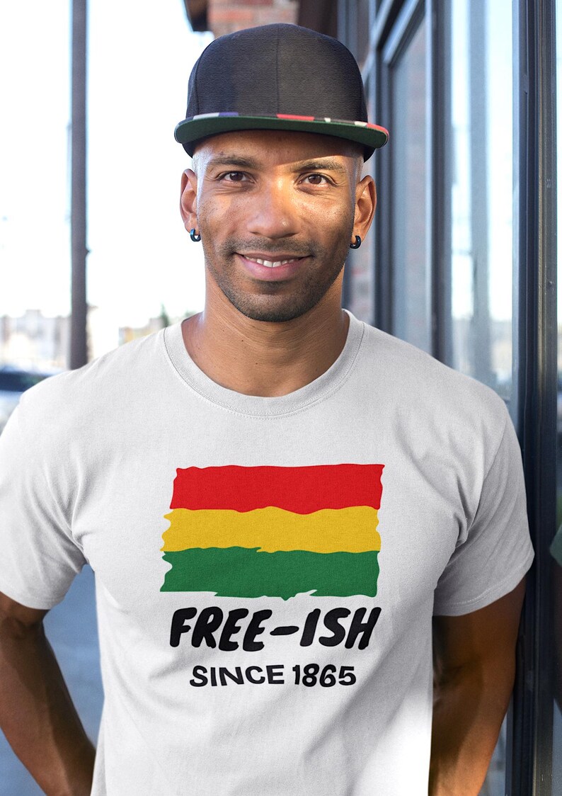 Op de afbeelding: Een wit T-shirt met korte mouwen en een afbeelding van een rood, geel en groen horizontaal gestreepte vlag met een verweerde textuur. Onder de vlag staat de tekst "FREE-ISH" in dikke zwarte letters, gevolgd door "SINCE 1865" in kleinere zwarte letters.