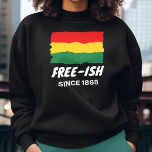 Op de afbeelding: Een zwart sweatshirt met lange mouwen en een afbeelding van een rechthoekige vlag in rood, geel en groen met een rafelige rand. Onder de vlag staat in witte, vette letters "FREE-ISH", gevolgd door de kleinere witte tekst "SINCE 1865".