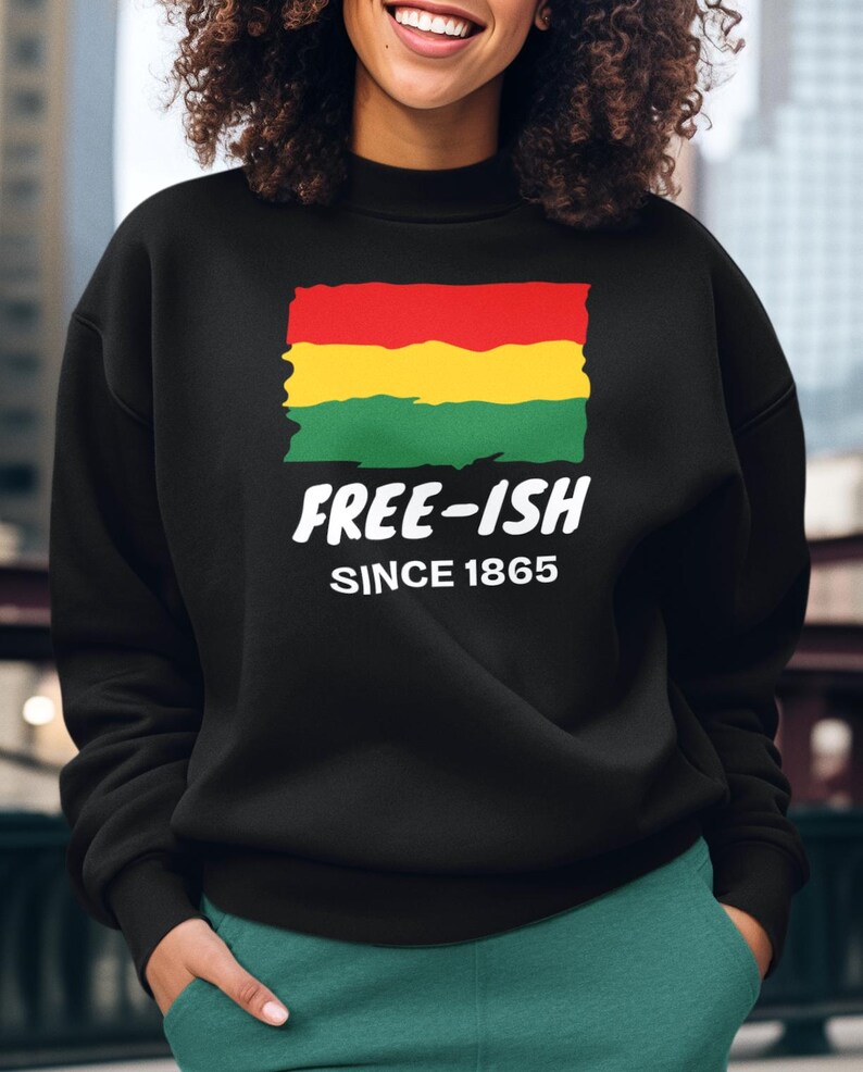 Op de afbeelding: Een zwart sweatshirt met lange mouwen en een afbeelding van een rechthoekige vlag in rood, geel en groen met een rafelige rand. Onder de vlag staat in witte, vette letters "FREE-ISH", gevolgd door de kleinere witte tekst "SINCE 1865".